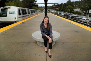 Susan Shaheen - in a navy patterned blouse and black pants - sits on a circular bench on the platform at the Orinda BART station in Orinda, California.