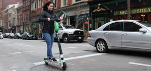 A woman in a black jacket rides a green Lime scooter on a street