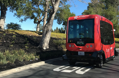 Image of a small, red boxy automated vehicle in front of some trees and shrubbery on a road